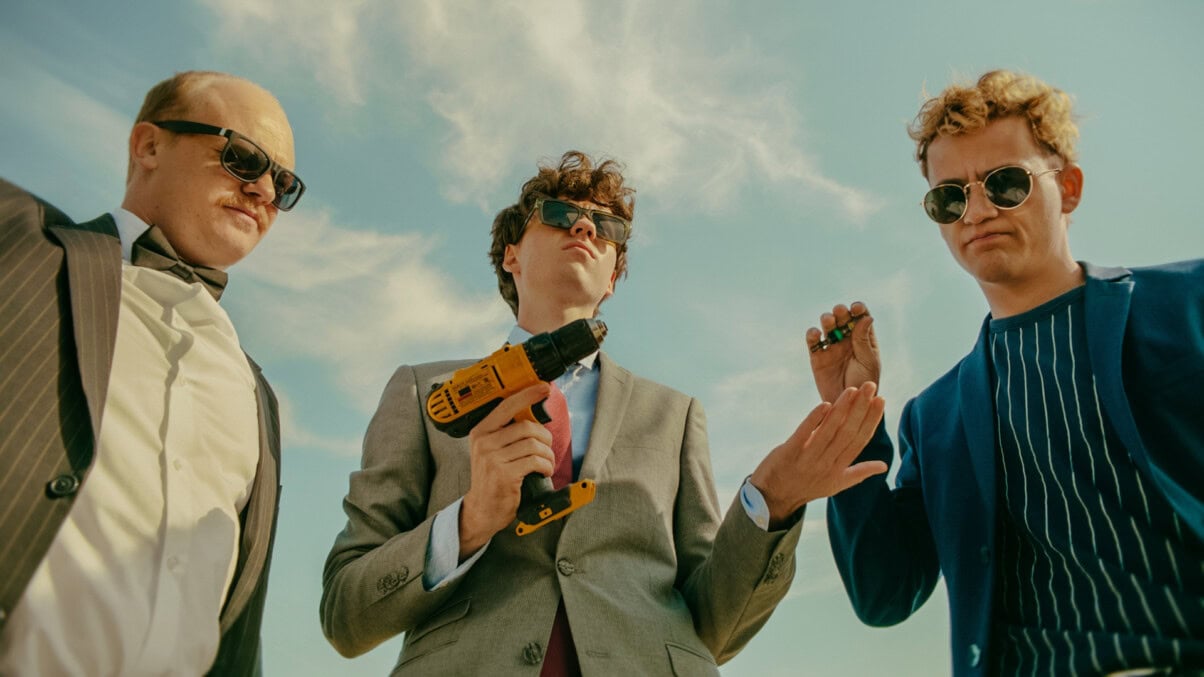 TV & Streaming Tipps Oktober: Drei Männer unter freiem Himmel in Anzügen und einem Akkuschrauber in der Hand.