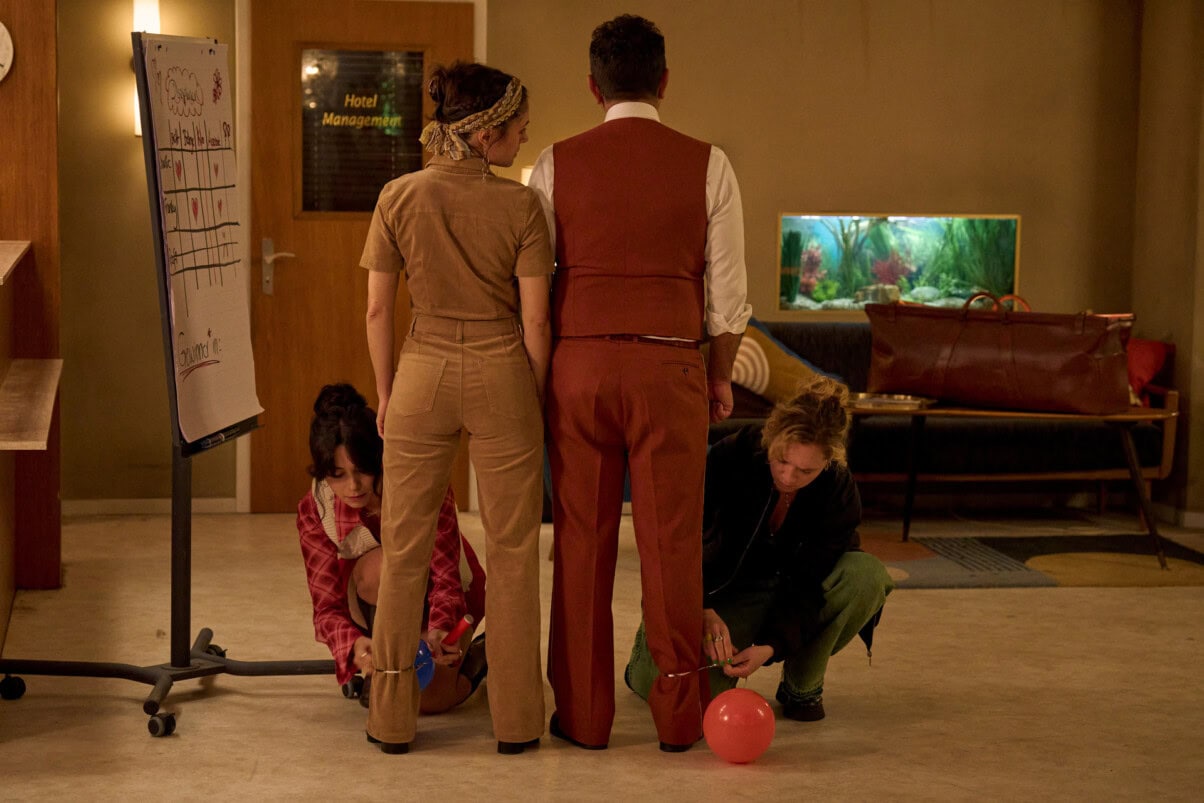 TV & Streaming Tipps im Juni "Nighties - Die Nacht mit dem Ring": Charles (Adnan Maral) und Franca (Mariananda Schempp) stehen in der Lobby eines Hotels mit dem Rücken zum Betrachter, während Millie (Tamara Romera Ginés) und Adriana (Stella Goritzki) vor ihnen knien und Luftballons an die Beine binden.