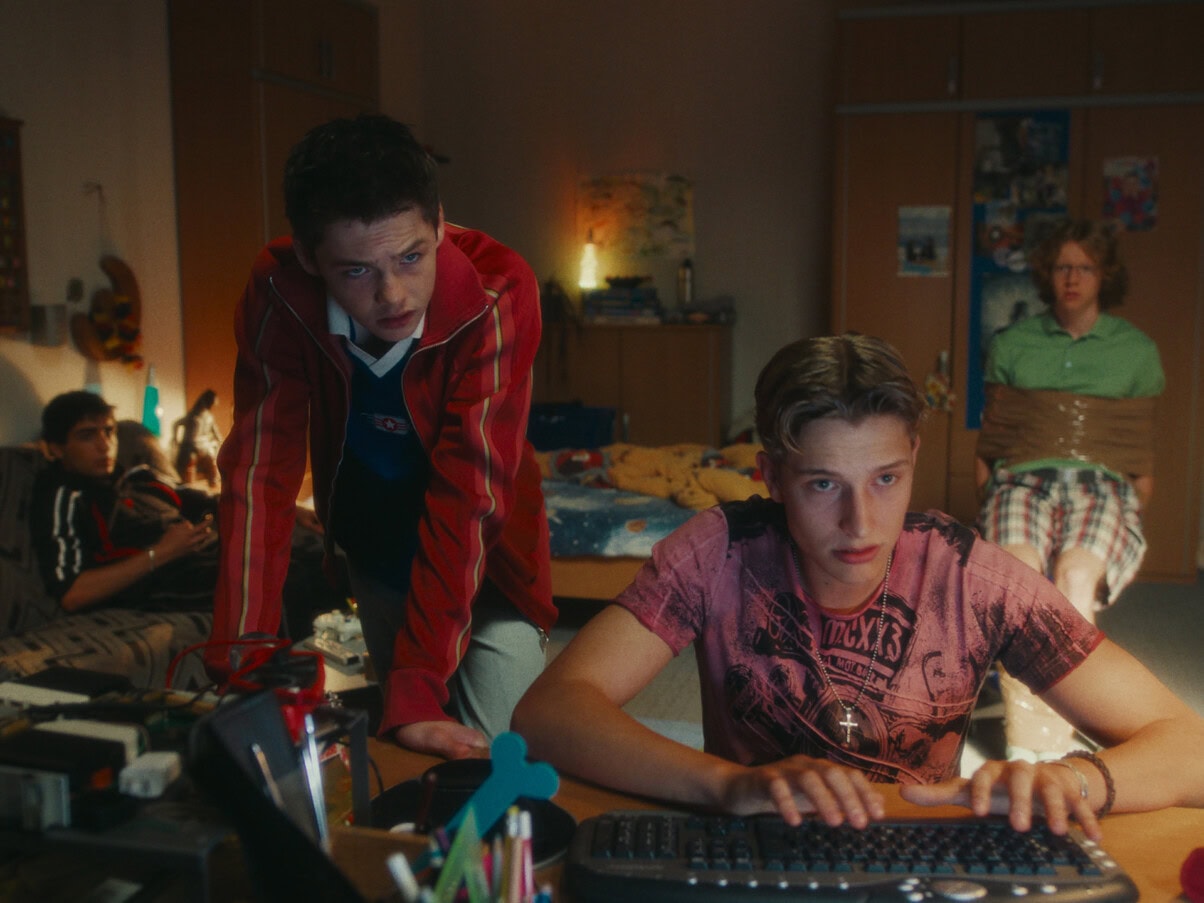 TV & Streaming Tipps im August 2025: In einem Kinderzimmer befinden sich vier Jungs im Teenager-Alter. Im Vordergrund sitzt einer am Schreibtisch an einer Tastatur, der zweite Lehnt über dem Schreibtisch und sieht in dieselbe Richtung wie der erste - vermutlich blicken beide in einen Computerbildschirm. Im Hintergrund sitzen zwei weitere Jungen - einer ist mit Paketklebeband gefesselt auf einem Stuhl, der zweite sitzt auf einer Couch - auch sie blicken beide Richtung Bildschirm.