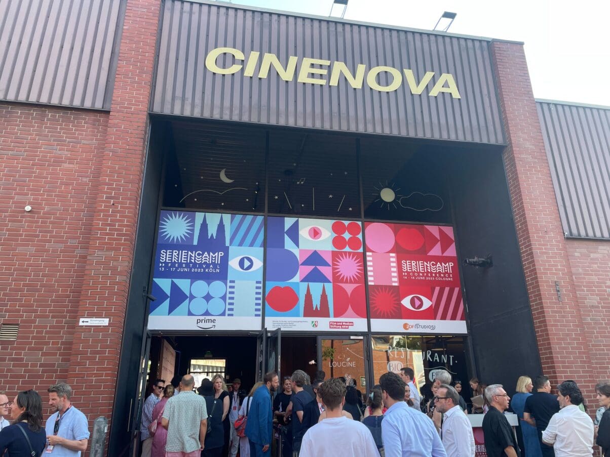 Seriencamp 2023 Opening (c) Medien.Bayern GmbH Das Cinenova Kino in Köln Ehrenfeld von Außen. Der Eingang ist mit dem blau-roten Design des Seriencamp Festivals & Seriencamp Conference gebranded. Vor dem Eingang stehen einige Leute und sprechen miteinander.