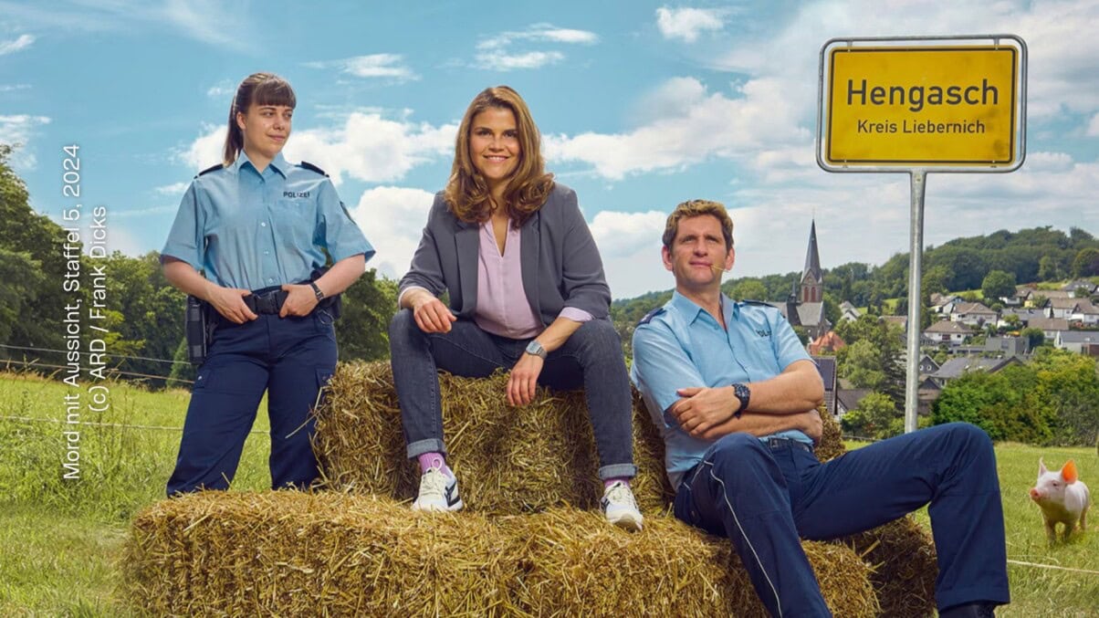 Mord mit Aussicht-Staffel5_c_ARD_FrankDicks Aus: Mord mit Aussicht – Staffel 5. Die Kommissarin sitzt zusammen mit einem in Uniform gekleideten Polizisten auf einem Stroh-Ballen. Daneben steht eine weitere Polizistin in Uniform. Die Kommissarin trägt einen Blazer und legere Kleidung - Bluse, Jeans, Sneaker. Sie blicken in die Kamera. Daneben ist ein Orsschild mit dem Text "Hengasch - Kreis Liebernich" zu sehen. Im Hintergrund sind viele Häuser und eine Kirche aus einem Dorf zu sehen. Schräg hinter den drei Personen ist ein kleines Schwein.
