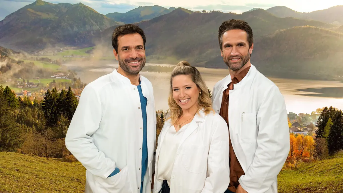 Die Landarztpraxis - Staffel 3_c_Joyn-Julia Feldhagen Die Hauptfiguren stehen in der Mittel. EIne Frau und zwei Männer. Alle tragen Arztkittel. Im Hintergrund ist eine wunderschöne Landschaft im Sonnenlicht zu sehen - mit Bergen, einem See und einer grünen Wiese.