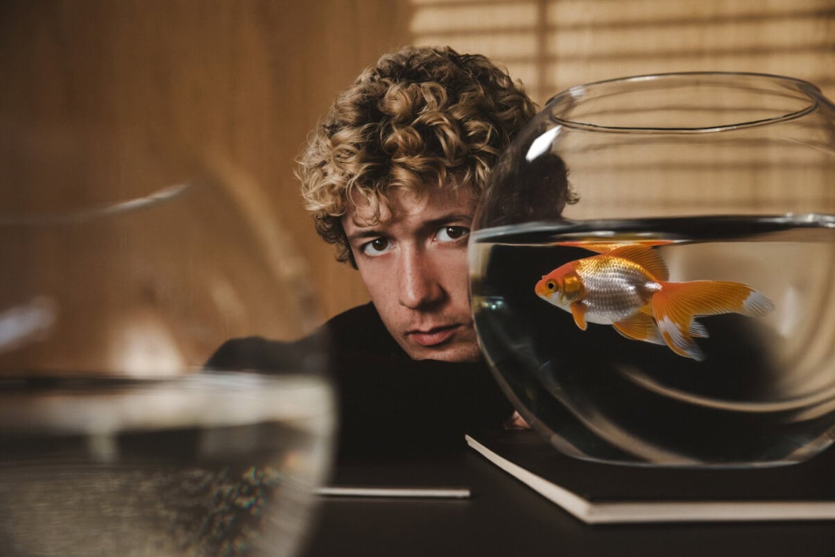 WTF_is_Jule_c_ZDF TV & Streaming Tipps Oktober: Mann auf einem Tisch gelehnt, teilweise durch ein Goldfischglas aufgenommen