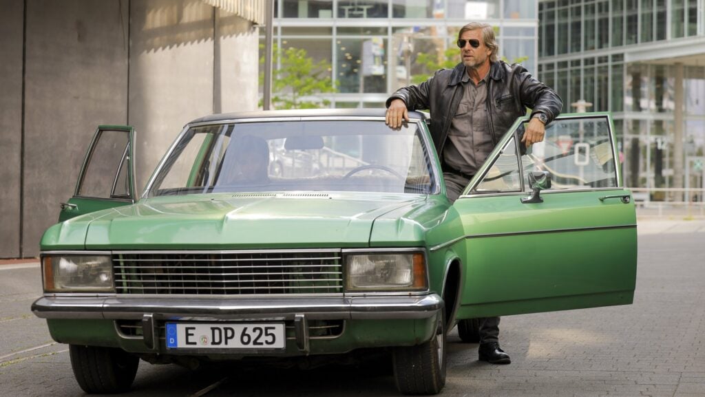 Der_letzte_Bulle_c_Joyn_Tom_Trambow TV & Streaming Tipps November: Ein Mann in Lederjacke und Sonnenbrille lehnt lässig an der geöffneten Tür eines grünen Oldtimers, der vor einem modernen Gebäude steht.