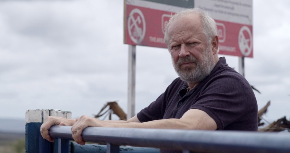 Verschollen_c_SWR_Diwafilm TV & Streaming Tipps November: Ein älterer Mann mit grauem Bart lehnt nachdenklich an einem blauen Geländer im Freien, hinter ihm sind Schilder und eine weite Landschaft zu sehen.