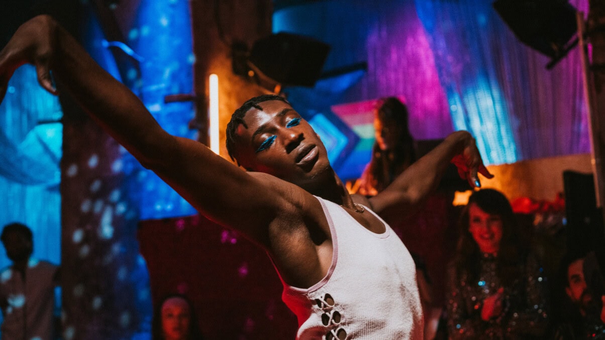 House_of_Bellevue_c_Daniel_LWOWSKI_ZDF TV & Streaming Tipps Dezember 2025: Eine Person mit blauem Augen-Make-up tanzt in einem Club mit weit ausgebreiteten Armen, trägt ein weißes Top und ist von buntem Neonlicht sowie unscharfen Personen im Hintergrund umgeben.