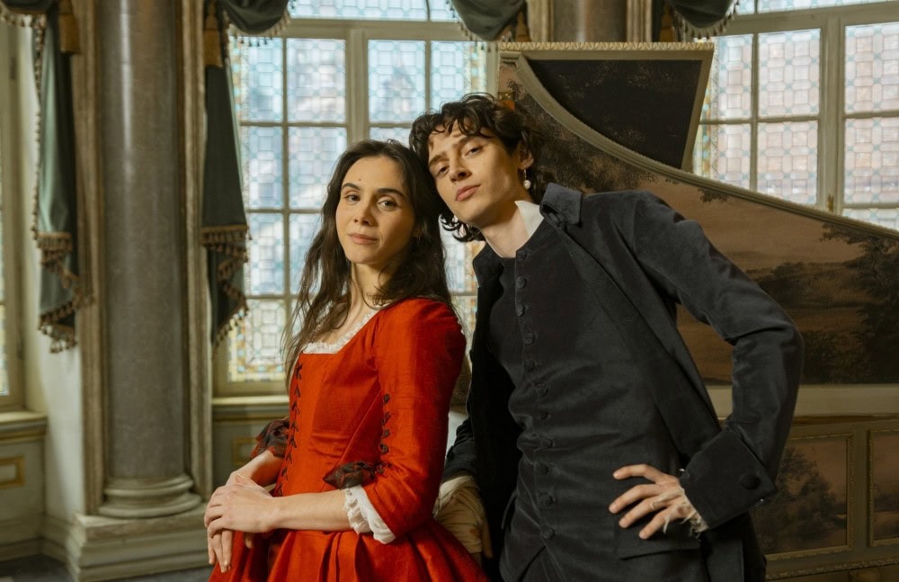 mozart_mozart_c_armands_virbulis_story_house_pictures TV & Streaming Tipps Dezember 2025: Zwei junge Erwachsene in historischer Kleidung – sie im roten Kleid, er im dunklen Anzug – lehnen selbstbewusst an einem Cembalo in einem prunkvollen Raum mit großen Fenstern.