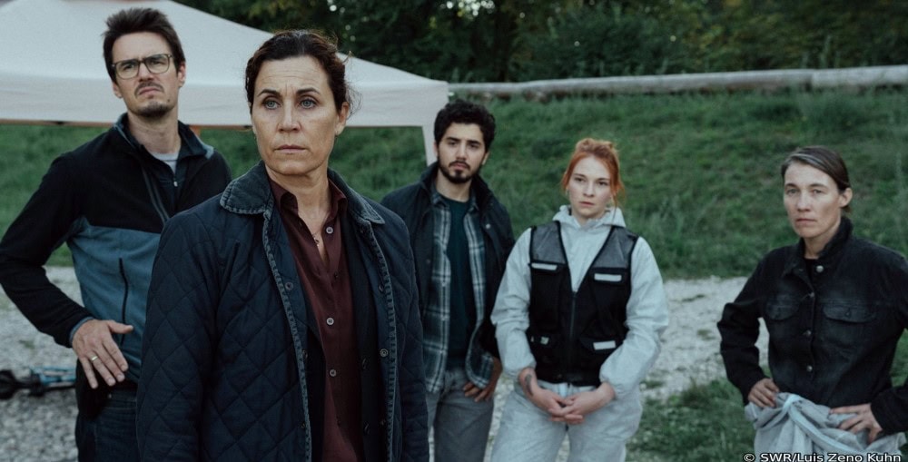 Spuren @SWR_Luis Zeno Kuhn Fünf Personen stehen in einer grünen Landschaft. Zwei der drei Frrauen tragen eine schusssichere Weste. Die Frau, die keine Weste trägt, steht im Vordergrund. Alle Personen schauen knapp an der Kamera vorbei. Die Stimmung wirkt gedrückt. Es ist Dämmerung.