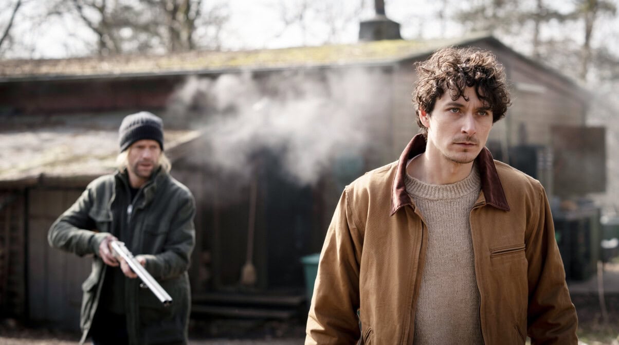 „Das zweite Attentat" in der ARD Mediathek. (c) Bild: ARD / WDR / ARD Degeto /Thomas Kost Zwei Männer stehen in einer trüben naturbelassenen Landschaft. Hinter ihnen steht ein Holzhaus. Der Mann rechts schaut bestimmt in die Ferne. Hinter ihm steht ein Mann mit einer Mütze mit einem Gewehr. Sein Blick ist offen, es wirkt, als hätte er gare einen Schuss losgelassen, da zwischen den beiden Männern eine Rauchwolke zu erkennen ist. Das Gewehr ist leicht gesenkt, die Stimmung wirkt angespannt.