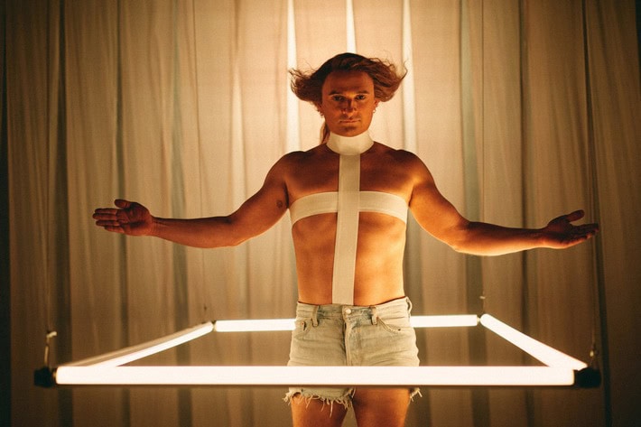 „Messiah Superstar" auf Joyn. (c) Joyn Ein Mann steht in einem viereckigen Rahmen, bestehend aus LED-Stäben. Der Mann trägt eine kurze Jeans-Short und kein Oberteil. Stattdessen hat er ein Kreuz über seine Brust mit weißem Klebeband geklebt sowie eine Art Rollkragen. Seine Arme sind ausgebreitet. Sein Blick ist starr nach vorne gerichtet und seine Haare sind an den Seiten zauselig nach oben gestylt.