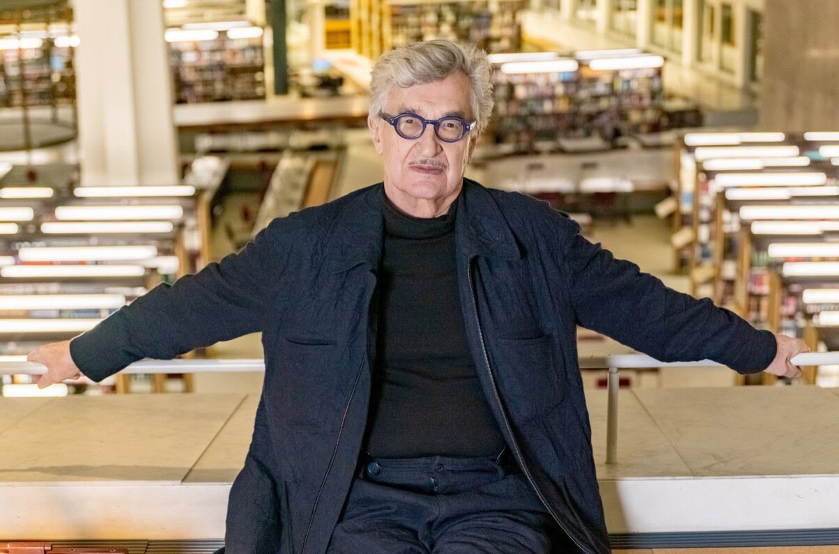 Wim Wenders: Der ewig Suchende TV & Streaming Tipps im August 2025: Regisseur blickt direkt in die Kamera. Er lehnt mit ausgestreckten Armen und seinem Rücken an einem Geländer. Der Hintergrund ist hell, aber unscharf. Vermutlich steht er im oberen Stockwerk einer Bibliothek. Er selbst trägt dunkbelblaue Kleidung.