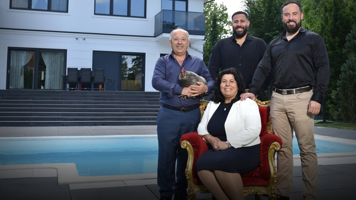 „Sons of Neukölln" (c) ZDF / André Kowalski TV & Streaming Tipps im August 2025: Vier Personen stehen etwas rechts im Zentrum des Bildes, alle lächeln in die Kamera. Zwei Männer mittleren Alters in schwarzen Hemd und hellbrauner Hose, eine ältere Frau in schwarzem Etuikleid und weißem Blazer die in einem eleganten Sessel sitzt und ein älterer Mann, blau gekleidet, der ein Huhn in seinen Händen hält. IM Hintergrund ist ein großes Haus sowie ein Pool zu sehen.