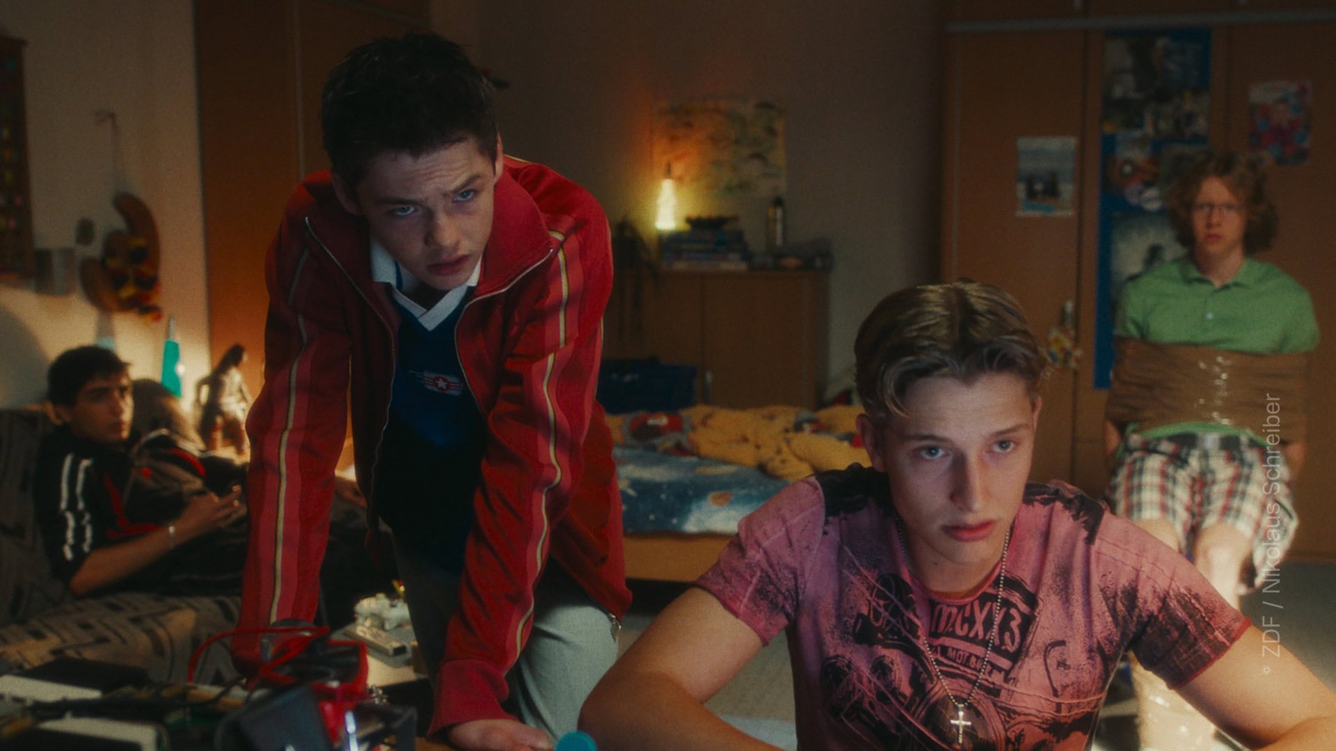 TV & Streaming Tipps im August 2025: In einem Kinderzimmer befinden sich vier Jungs im Teenager-Alter. Im Vordergrund sitzt einer am Schreibtisch an einer Tastatur, der zweite Lehnt über dem Schreibtisch und sieht in dieselbe Richtung wie der erste - vermutlich blicken beide in einen Computerbildschirm. Im Hintergrund sitzen zwei weitere Jungen - einer ist mit Paketklebeband gefesselt auf einem Stuhl, der zweite sitzt auf einer Couch - auch sie blicken beide Richtung Bildschirm.