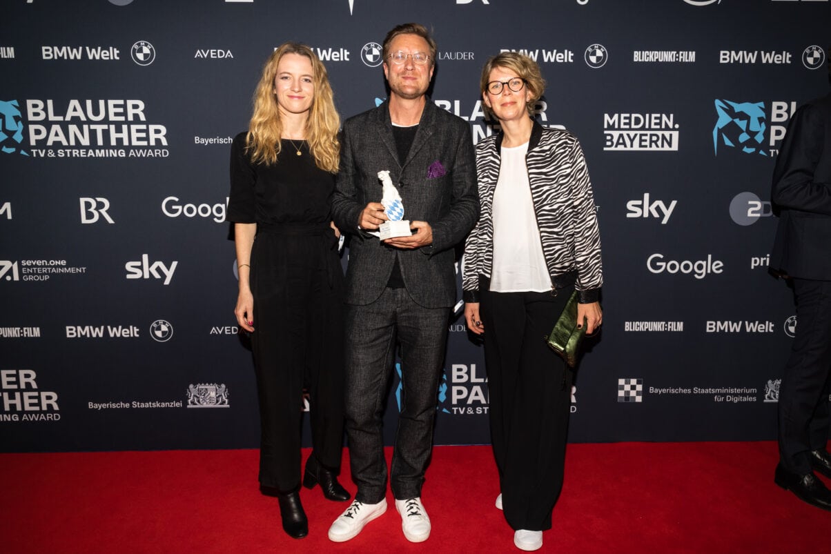 Preisträger Carl Gierstorfer mit Mareike Müller (Idee) und Antje Böhmert (Produktion) (c) Medien.Bayern Johannes Müller Preisträger Carl Gierstorfer (in der Mitte) mit Mareike Müller (Idee) und Antje Böhmert (Produktion) vor der Pressewand des Blauen Panthers 2022