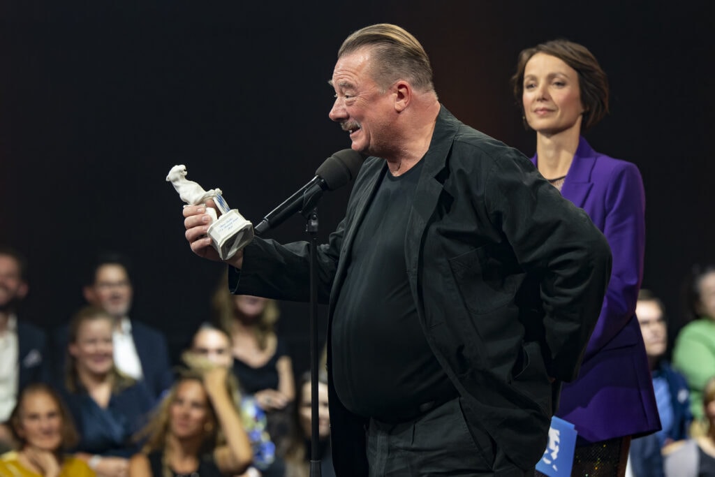 Peter Kurth (c) Medien.Bayern _ Ralf Wilschewski Peter Kurth mit dem Preis in der Hand, vor dem Mikrofon. Er hält gerade seine Dankesrede in der Show 2022.