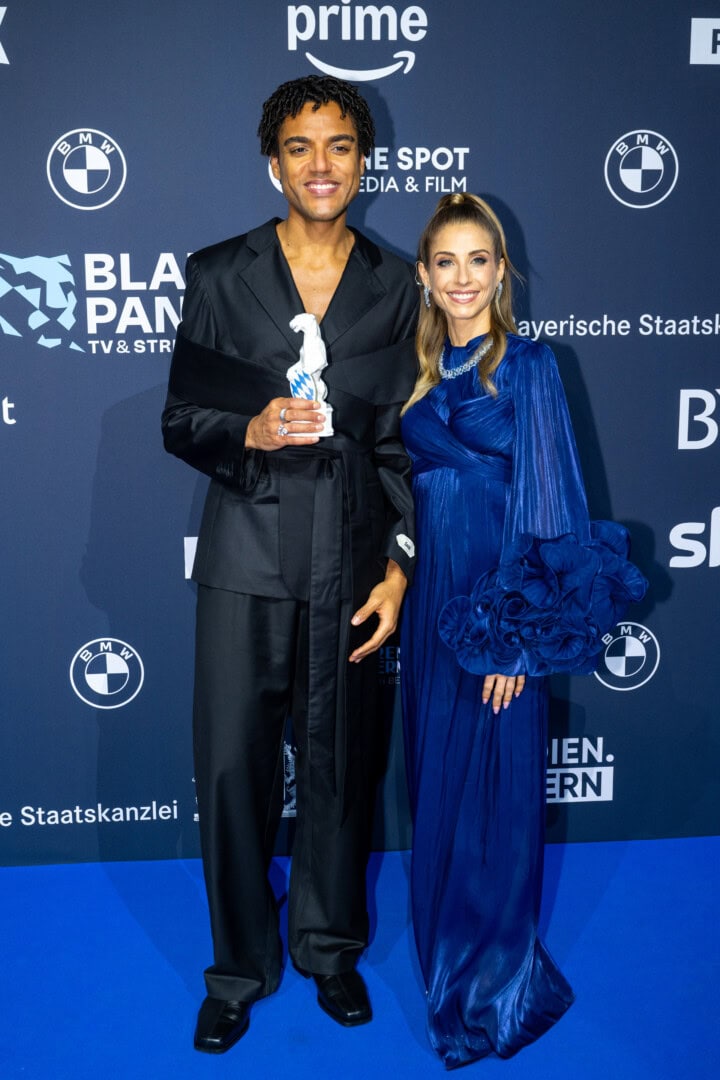 Lamin Leroy Gibba (l.) freut sich mit Moderatorin Cathy Hummels (r.) über den Blauen Panther 2025 in der Kategorie Fiktion als Hauptdarsteller, Creator, Ko-Produzent und Headautor von „Schwarze Früchte“ (Jünglinge Film & Studio Zentral / ARD Degeto Film für die ARD).