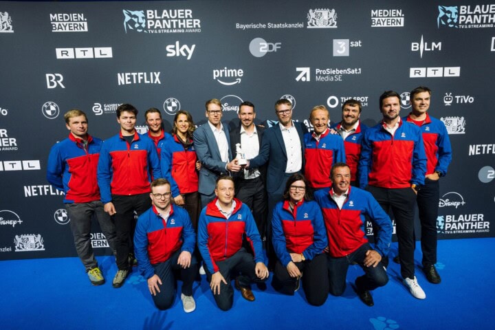 Franz Hinterbrandner (Mi.-l.) und Max Reichel (Mi.-r.) erhalten den „Blauer Panther – TV & Streaming Award“ 2026 für das „Beste Factual-Entertainment-Format“ für Produktion und Kamera in „In höchster Not – Bergretter im Einsatz“ (Timeline Production im Auftrag des Bayerischen Rundfunks für die ARD Mediathek) ausgezeichnet. Laudator Felix Neureuther (Mi.) und 12 Bergretter freuen sich mit den Gewinnern.